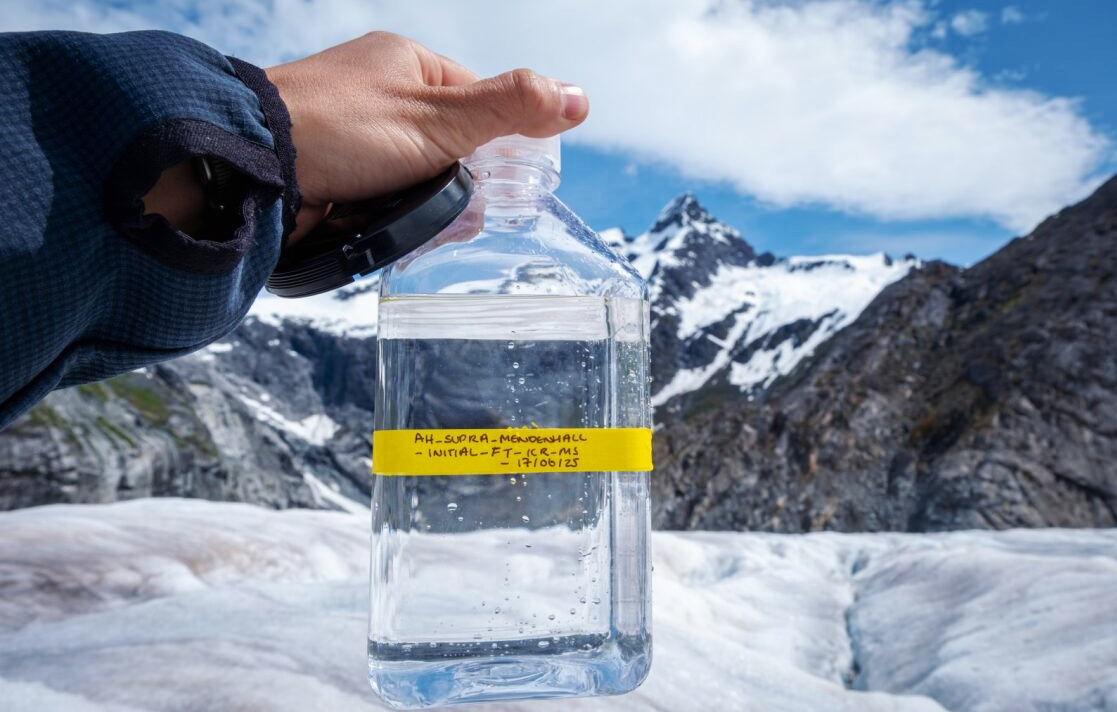 A labeled bottle containing glacier water sample with mountains and a glacier in the background